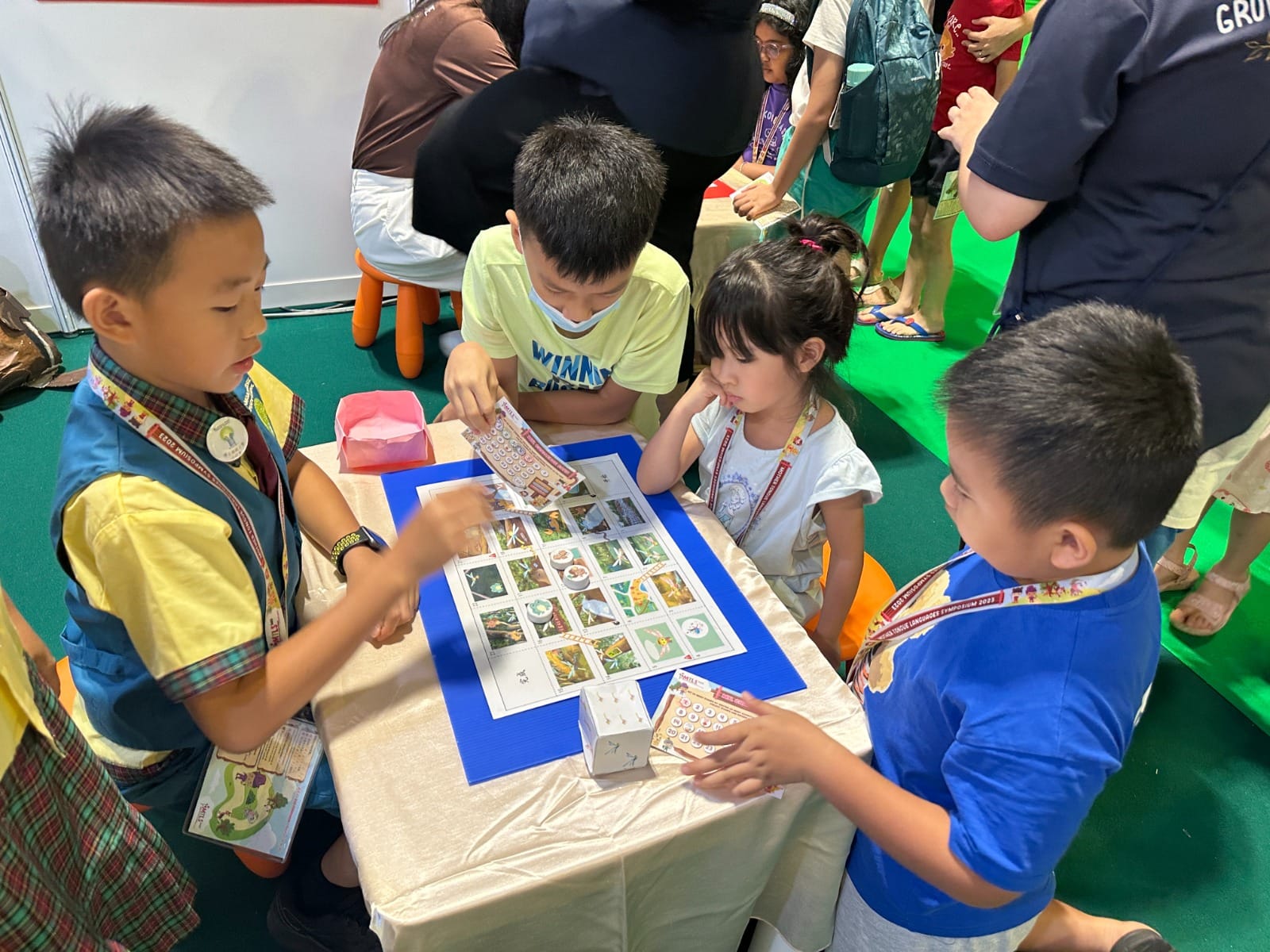 Four young children gather around a table playing a nature-themed card and board game at an indoor event.